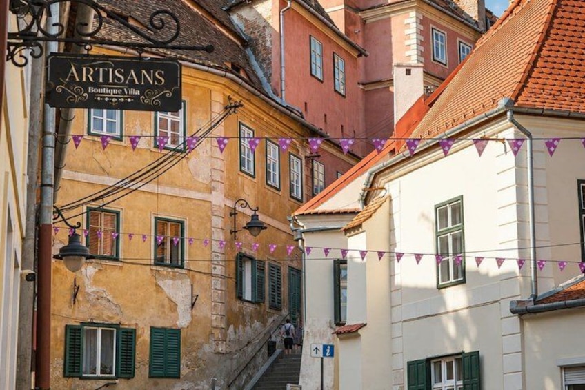 A narrow street with buildings, a sign for 'ARTISANS Boutique Villa,' and purple bunting. 1