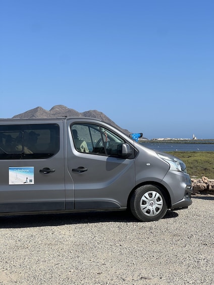 A silver car parked on a gravel surface with a coastal landscape in the background. 2