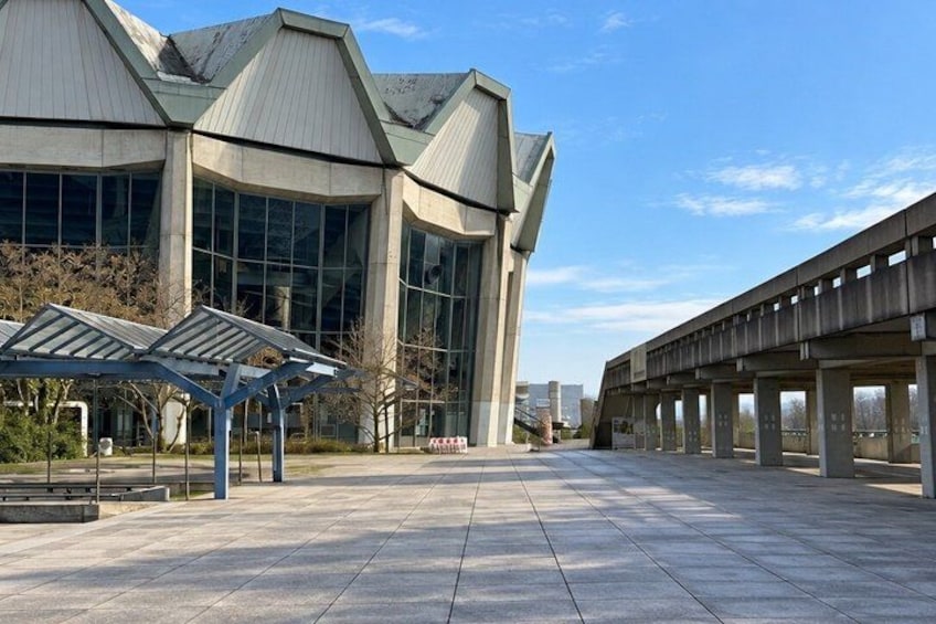 A modern building with a unique roof structure and large windows, a covered walkway, and a bridge in the background. 5