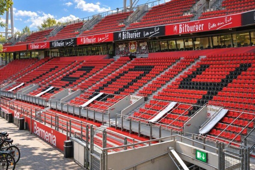 Un estadio con asientos rojos y negros, un anuncio destacado de Bitburger y una señal de salida visible. 5