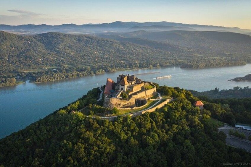 A castle on a hill overlooking a river, surrounded by forested hills. 1