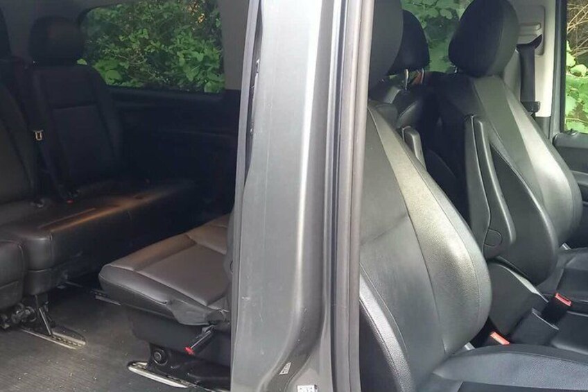 A vehicle interior with black leather seats, a visible seatbelt, and a window with greenery outside. 2