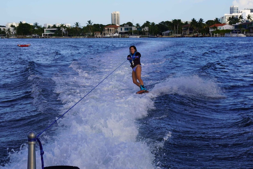 Eine Person beim Wakeboarden auf einem Gewässer mit Wellen, ein Boot im Hintergrund und eine Küstenstadt mit Gebäuden und Palmen. 1
