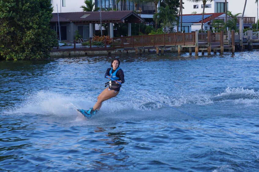 Eine Person treibt auf einem Wakeboard über das Wasser, im Hintergrund ein Holsteig und Häuser. 5