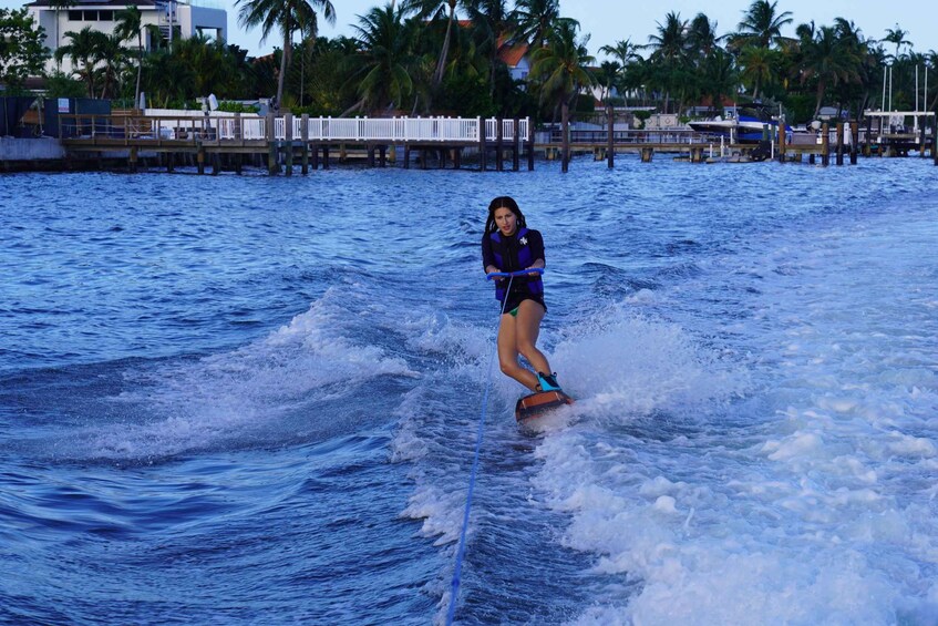 Eine Person treibt auf einem Wakeboard über ein Gewässer, im Hintergrund Palmen und Gebäude. 4