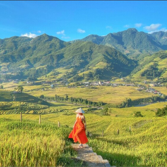 A person in an orange dress walking on a path through a green field with terraced hills and a village in the background. 1