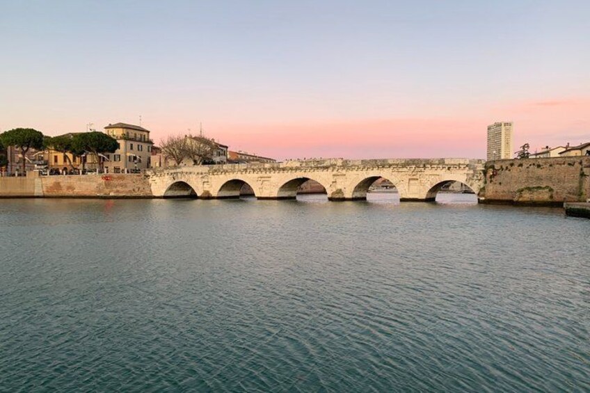 A stone bridge over a calm river with buildings on either side. 3
