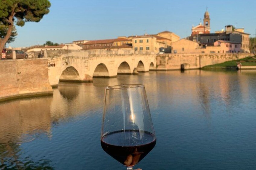 A glass of red wine in the foreground with a historic bridge and buildings in the background. 4