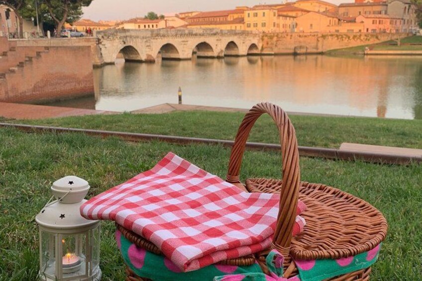 A picnic setup with a wicker basket, a checkered cloth, and a lantern on grass near a river with bridges and buildings in the background. 5