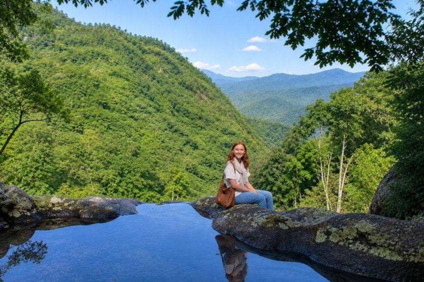 A person sitting on a rock by a clear blue pool surrounded by lush green hills. 1