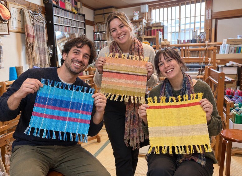 Drie mensen houden geweven textiel vast in een werkplaats met rekken vol wol en breigereedschap. 1