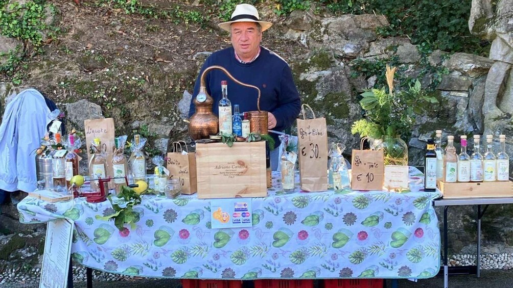 A person standing behind a table with various bottles and bags, possibly at a market or outdoor event. 5