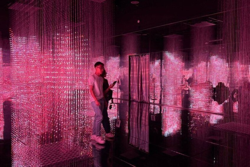 A person playing a guitar in a room with pink lighting and a large digital screen displaying abstract patterns. 1
