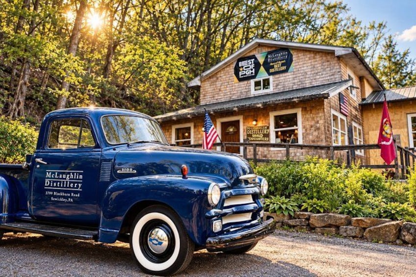 A vintage blue pickup truck parked in front of a distillery building with American flags. 1