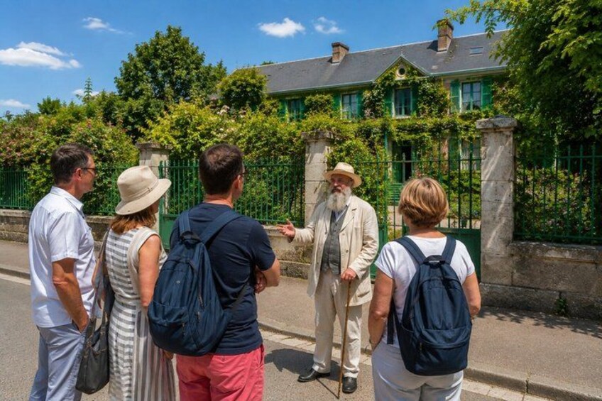 A group of people standing on a street, a green house with a fence, and trees in the background. 2