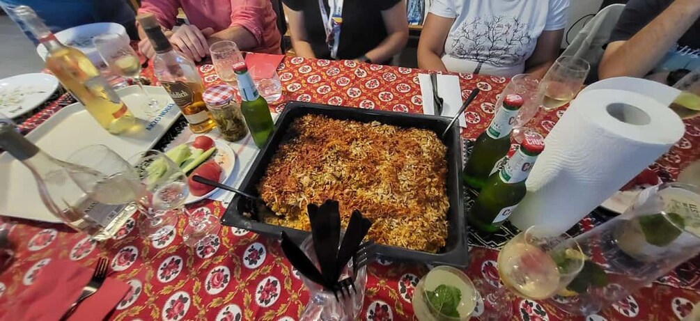 A dining table with a large dish of food, wine bottles, glasses, and a roll of paper towels. 3