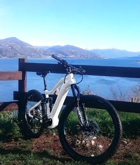 A white mountain bike parked on a grassy area with a wooden fence, overlooking a lake and mountains in the distance. 2