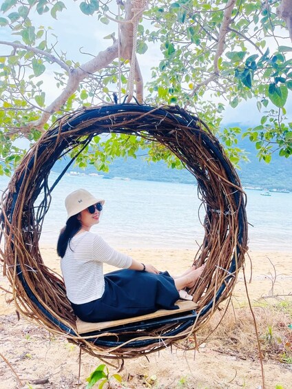 A person sitting in a circular wicker swing by a beach with a body of water in the background. 2