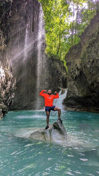 A person standing on a rock in a clear blue water body with a waterfall in the background. 1