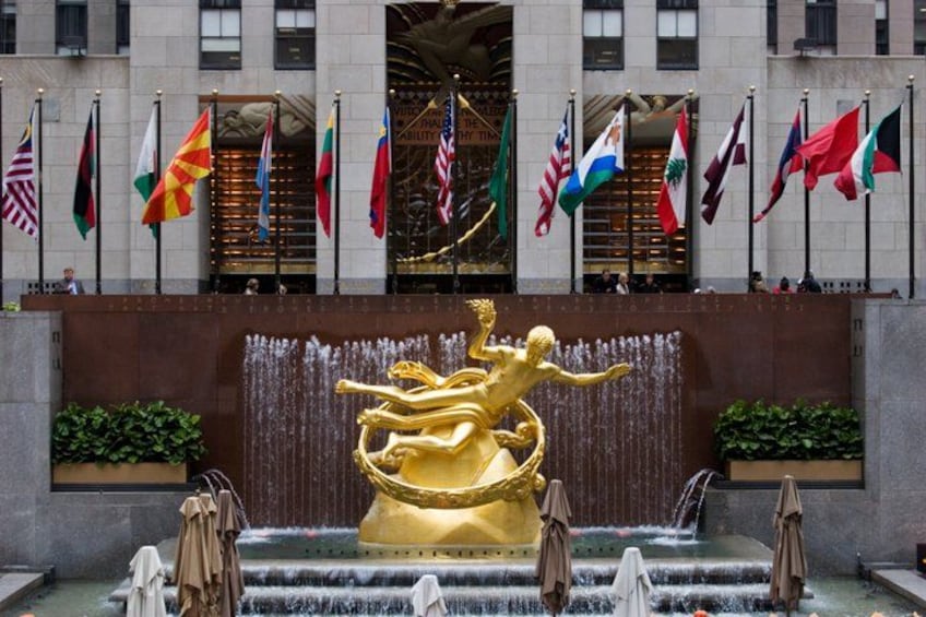 A golden statue of a figure in a dynamic pose, surrounded by a water feature with multiple fountains. Flags of various countries are displayed above the building. 1