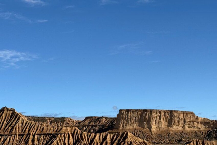 A vast, rugged landscape with layered rock formations under a clear blue sky. 2