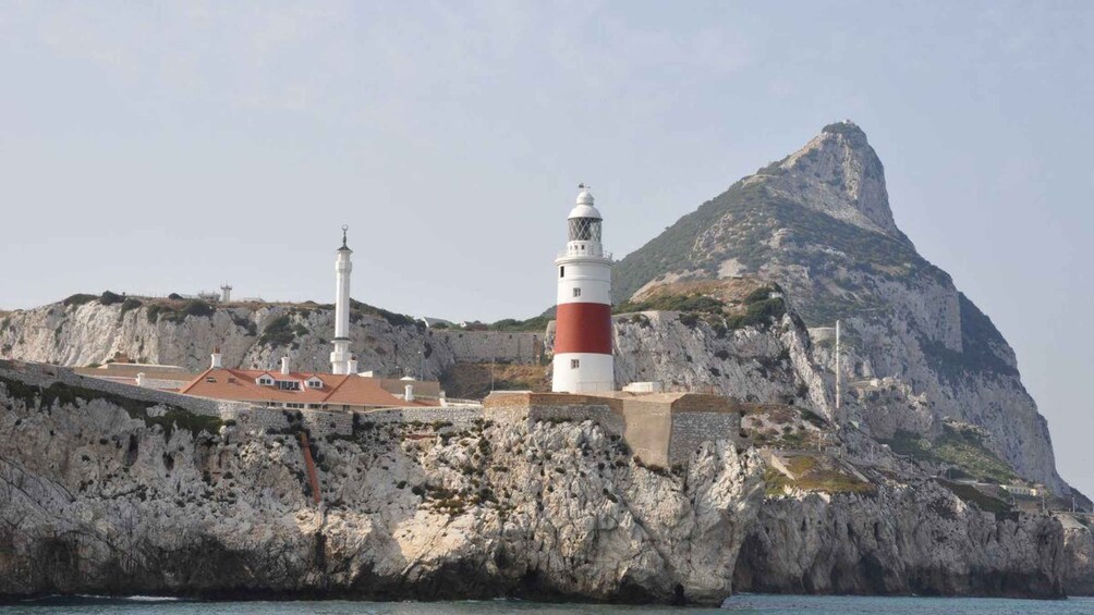 Un faro costero con una torre a rayas rojas y blancas, una torre blanca, y un acantilado rocoso con un pico destacado. 1