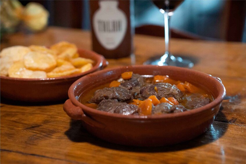 A bowl of beef stew with carrots and potatoes, a bottle of sauce, and a glass of wine on a wooden table. 4