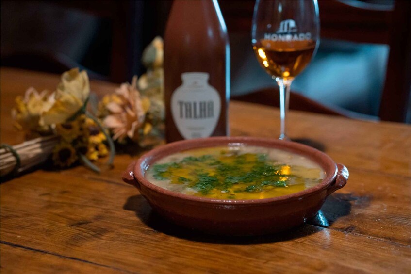 A bowl of soup with herbs on a wooden table, a bottle labeled 'TALIBO', and a glass of wine. 2