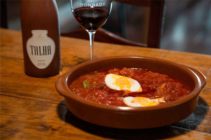 A bowl of stew with a poached egg on top, a bottle of Talha wine, and a glass of red wine on a wooden table. 5