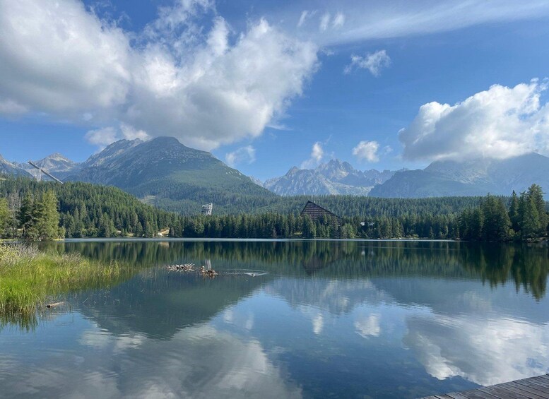 A tranquil lake with a wooden dock, surrounded by dense forests and towering mountains under a partly cloudy sky. 3