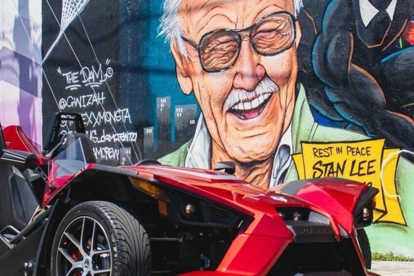 En rød sportsvogn parkeret foran en mural med teksten 'REST IN PEACE STAN LEE'. 3