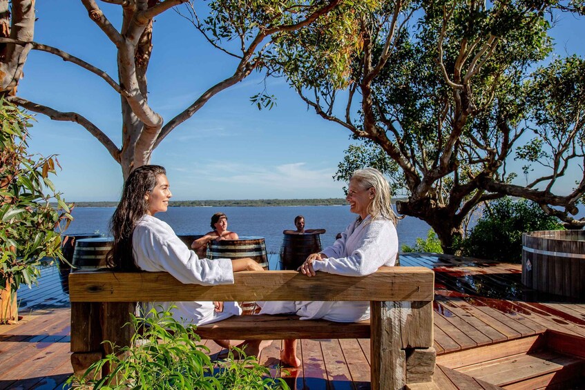 Three people sitting on a wooden bench by a lake, with trees and a wooden deck in the background. 2