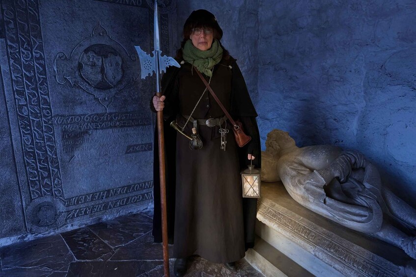 A person in historical costume holding a staff and a lantern, standing in front of a stone wall with carvings. 2