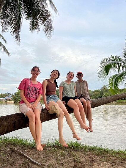 Four people sitting on a tree branch by a calm body of water, with a cloudy sky above. 2