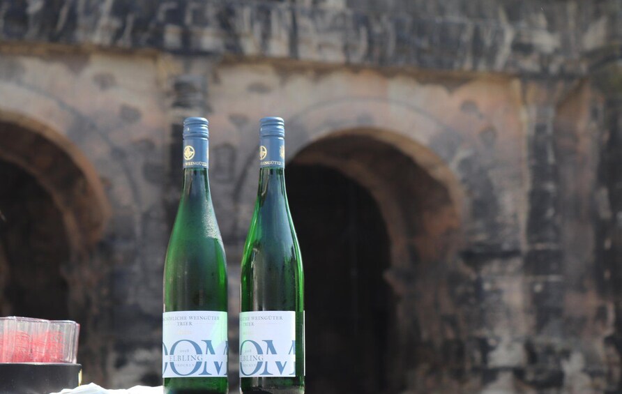 Two bottles of wine with labels in front of a stone building with arched doorways. 2