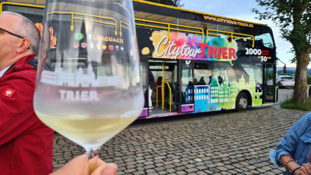 A person holding a wine glass with the word 'TRIER' on it, standing next to a colorful bus with 'CityTour Trier' written on it. 5