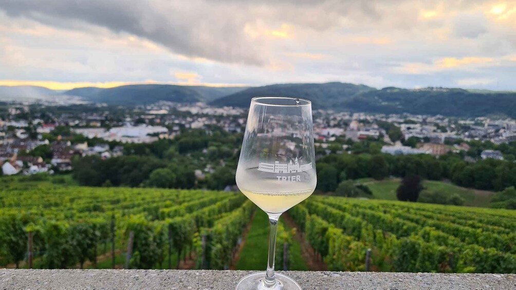 A wine glass with 'TRIER' engraved on it, set against a backdrop of a vineyard and a town. 1