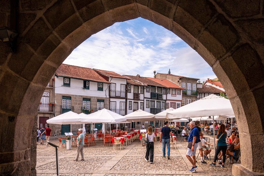Un arco de piedra con vista a una plaza adoquinada, con mesas al aire libre y edificios al fondo. 1