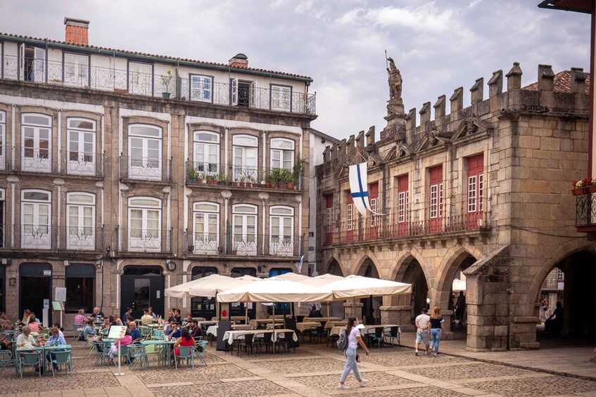 Una plaza histórica con mesas al aire libre, una bandera y arquitectura ornamentada. 3