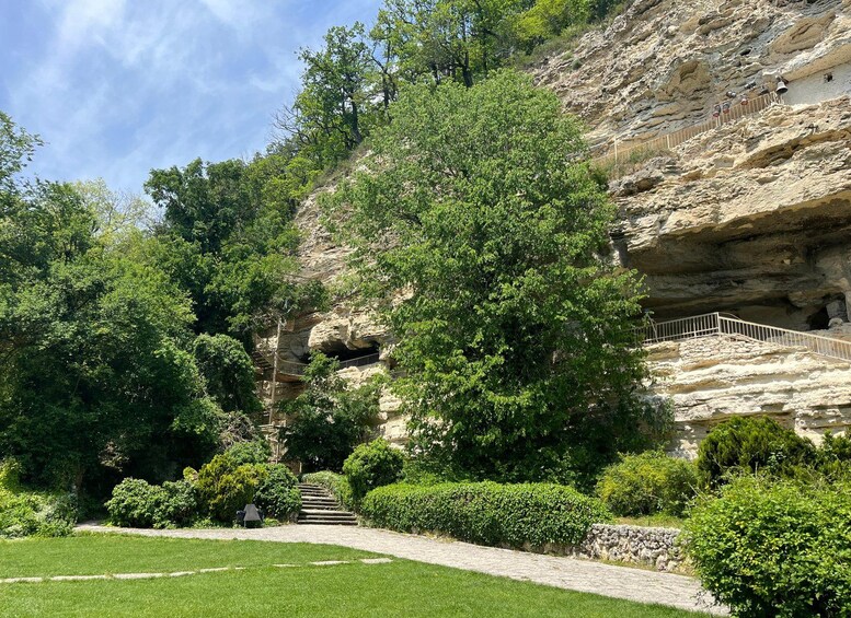 A rock formation with a staircase leading up to a cave entrance, surrounded by lush greenery and a well-maintained lawn. 5