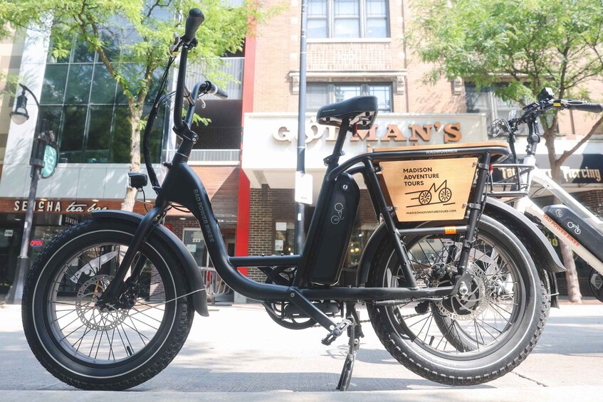 A black electric bike with a wooden box labeled 'Madison Adventure Tours' parked on a sidewalk. 3