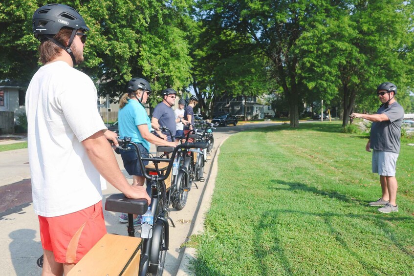 A group of people on bicycles lined up on a sidewalk, with a grassy area and trees in the background. 4