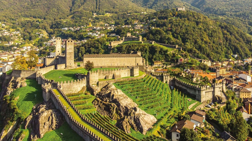 A historic walled city with a castle, terraced vineyards, and a mountainous backdrop. 1