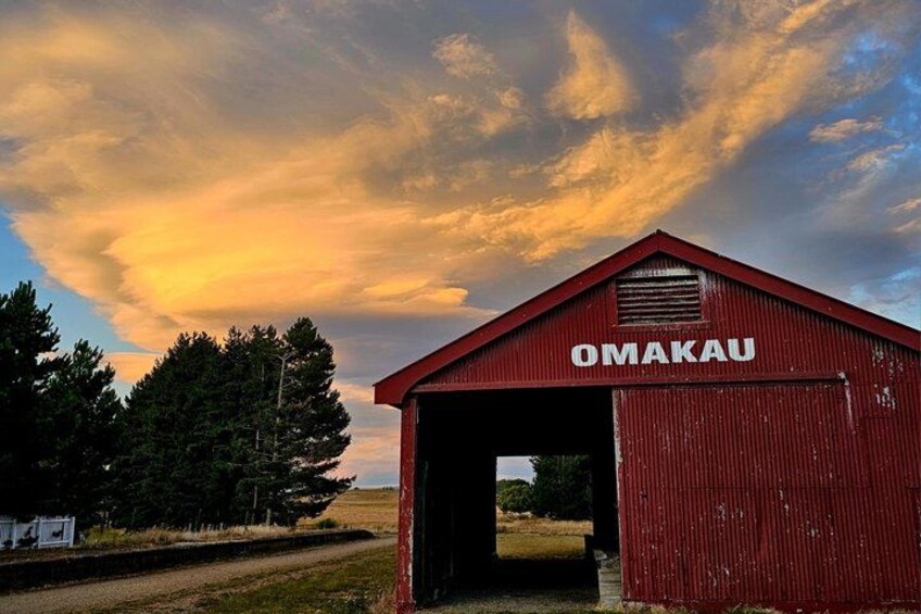 A red barn with the name 'OMAKAU' on a rural landscape with a cloudy sky. 3