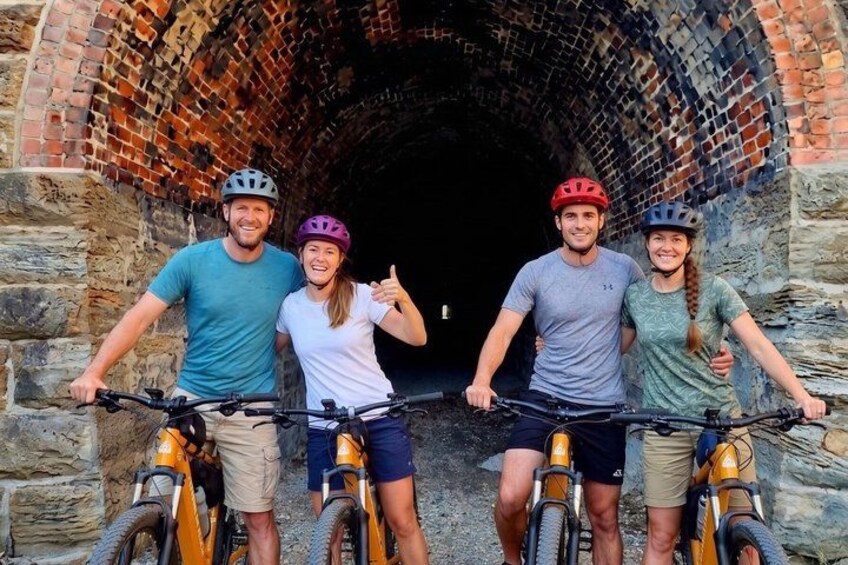 Four people on bicycles in front of a brick tunnel. 1