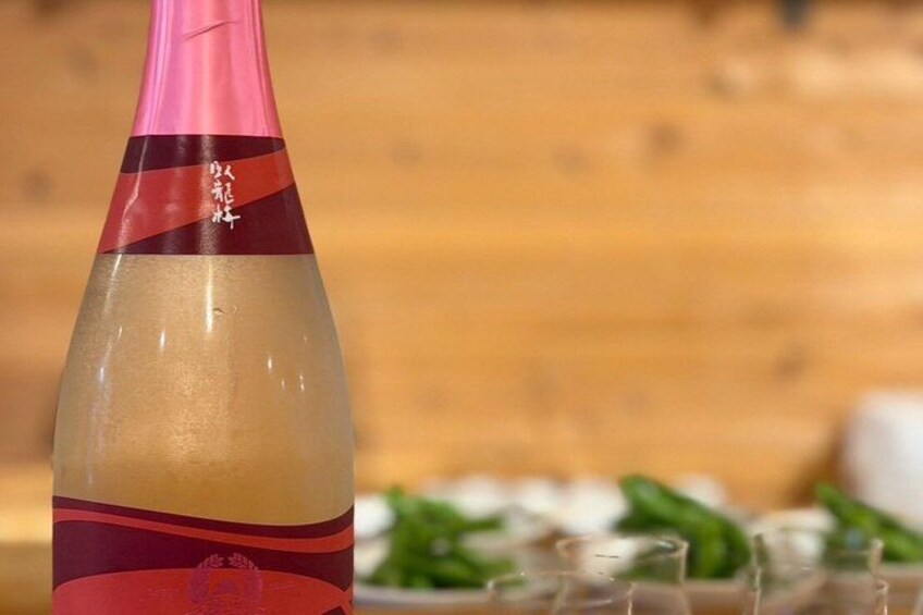 A bottle with a pink and red label, a wooden background, and a plate with green vegetables. 3