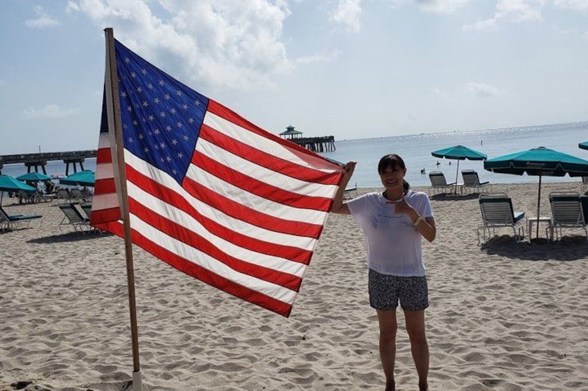 A person holding an American flag on a beach with umbrellas and lounge chairs in the background. 1