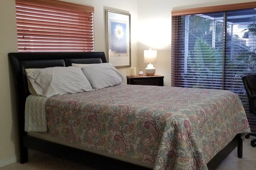 A bed with a patterned bedspread, a nightstand with a lamp, and a window with blinds. 5