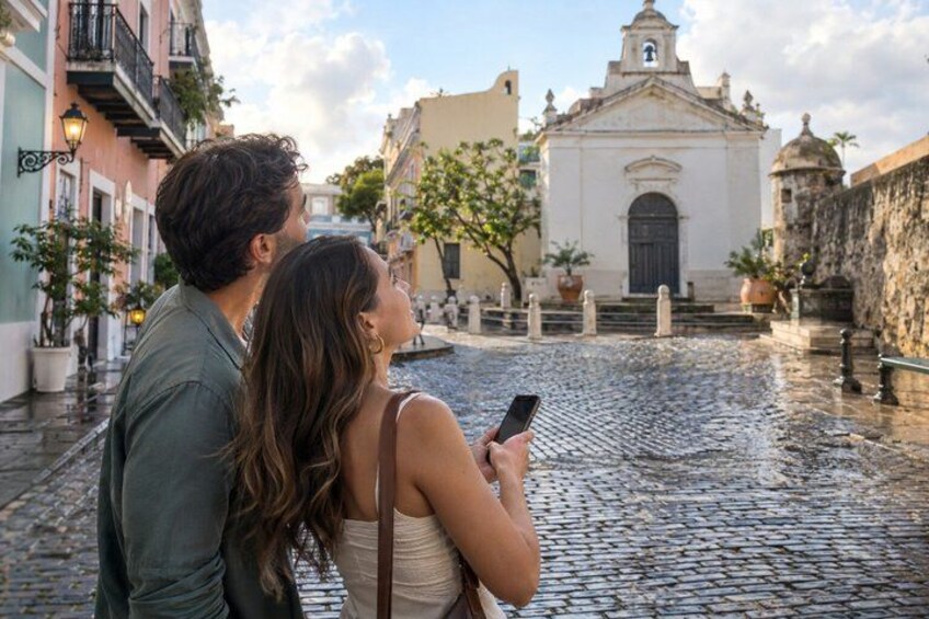 A couple walking on a cobblestone street, a church, and buildings with balconies. 4