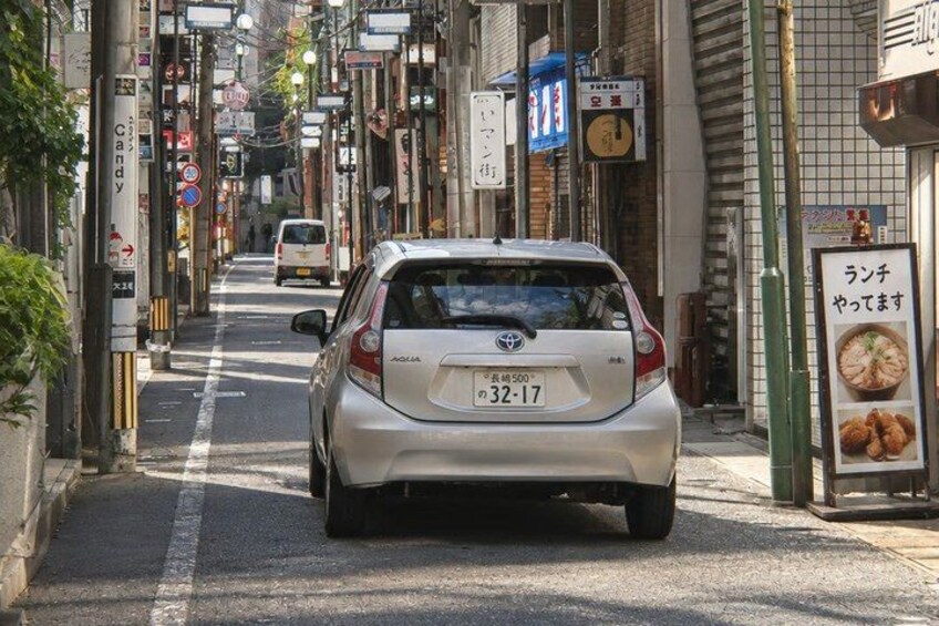 A narrow urban street with parked cars, storefronts, and various signs. 2